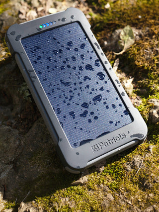 Patriot Power Cell-CX Portable Solar Power Bank charging in sunlight on wet mossy ground, water beading on the rugged solar panel of this off-grid emergency charger