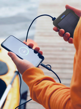 Person holding the Patriot Power Cell-CX Portable Solar Power Bank charging a phone to 84% outdoors by a kayak, off-grid emergency solar charger