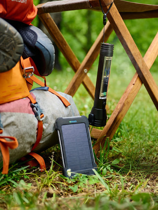 Patriot Power Cell-CX Portable Solar Power Bank propped against a backpack beside a hanging flashlight at a campsite, ready off-grid emergency solar charging power