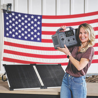 Woman holding the Patriot Power Generator 1200 by its handle beside a folding solar panel and American flag, a portable solar backup generator