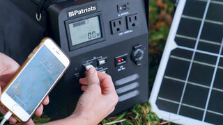 Patriot Power Generator 1800 portable power station on grass beside a folding solar panel, a hand plugging a smartphone into its USB port to charge, off-grid emergency backup power and portable solar