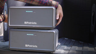 Two stacked Patriot Power Generator 2200X expansion battery packs in a truck bed with a folding solar panel as a man handles them, off-grid emergency backup power and portable solar