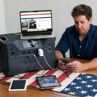 Patriot Power Solar Generator 2500X powering a laptop, tablets and phone on a dining table as a man works, portable solar generator for home emergency backup power during outages