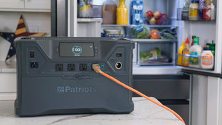 Patriot Power Solar Generator 2500X portable power station at 100% charge on a kitchen counter, an orange cord from its outlet powering a stocked fridge, off-grid emergency backup power for blackouts
