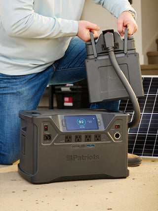 Man kneeling to connect an EXP expansion battery pack to the Patriot Power Solar Generator 2000X beside a solar panel, expanding off-grid emergency backup power capacity