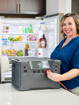 Patriot Power Solar Generator 2000X on a kitchen counter powering an open, stocked refrigerator as a smiling woman plugs in the cord for off-grid emergency backup power