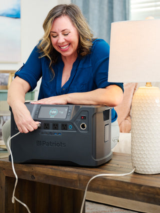 Patriot Power Solar Generator 2000X on a living room side table powering a lamp as a woman plugs it in, delivering portable solar off-grid emergency backup power indoors