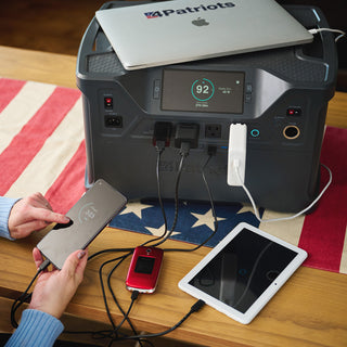 Patriot Power Solar Generator 2000X on an American flag runner charging a laptop, phone, tablet and flip phone, a portable solar generator for off-grid emergency backup power
