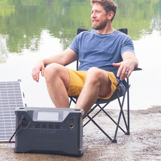 Patriot Power Solar Generator 2000X portable solar generator beside a man relaxing in a camp chair by a lake, recharging from a solar panel for off-grid backup power