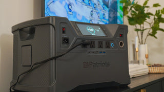 Patriot Power Solar Generator 2000X portable power station at 100% charge on a media console, its AC outlet powering a wall-mounted TV with a weather map, off-grid emergency backup power for blackouts