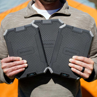 Patriot Power Voyager XL Solar Power Bank 3-pack held by a man outdoors by a tent, rugged portable solar chargers for off-grid camping and emergency power