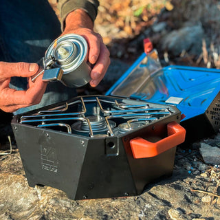 Man loading a fuel can into the RescueHeat Emergency Space Heater & Stove outdoors on rock, a portable off-grid cook stove and heater for emergency preparedness