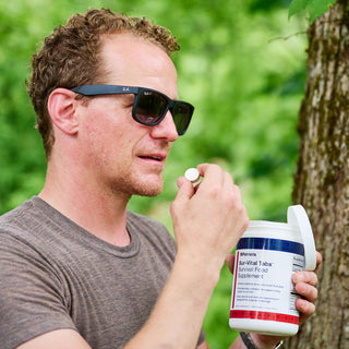 Man wearing sunglasses and a gray t-shirt holding a 4Patriots Sur-Vital Tabs container and taking a lemon-flavored survival food supplement tablet outdoors in a wooded setting