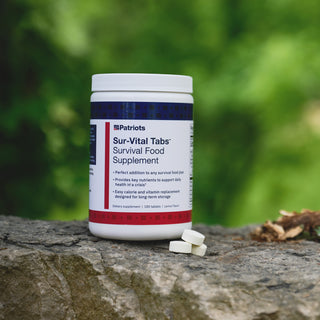  4Patriots Sur-Vital Tabs Survival Food Supplement 180-tablet container resting on a rock outdoors with two lemon-flavored tablets beside it and green foliage in the background