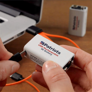 Hands holding a 4Patriots USB Rechargeable 9V battery being plugged in with an orange USB cable near a laptop, with a second battery in the background