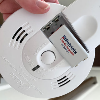 Hand inserting a 4Patriots USB Rechargeable 9V battery into the open compartment of a smoke and carbon monoxide alarm