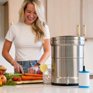 Patriot Pure Ultimate Water Filtration System & Nanomesh Filter on a kitchen counter as a woman chops fresh vegetables nearby with a glass of filtered water