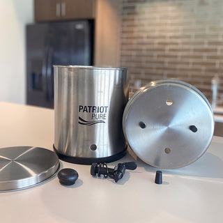 Patriot Pure Ultimate Water Filtration System Replacement Parts laid out on a kitchen counter, showing stainless chambers, base plate, spigot and stopper