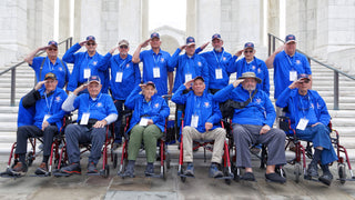 Group of senior military veterans in matching blue Honor Flight shirts and caps saluting on marble steps, several seated in wheelchairs, at a Washington memorial