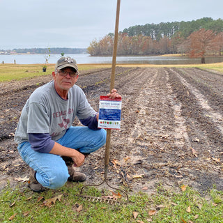 Man kneeling beside a freshly tilled garden plot holding a 4Patriots Victory Garden Seed Collection bag on a rake handle, with a lake and trees in the background