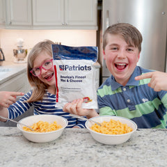 Two Kids holding an America's Finest Mac & Cheesepouch with some prepared Mac & Cheese on the counter.